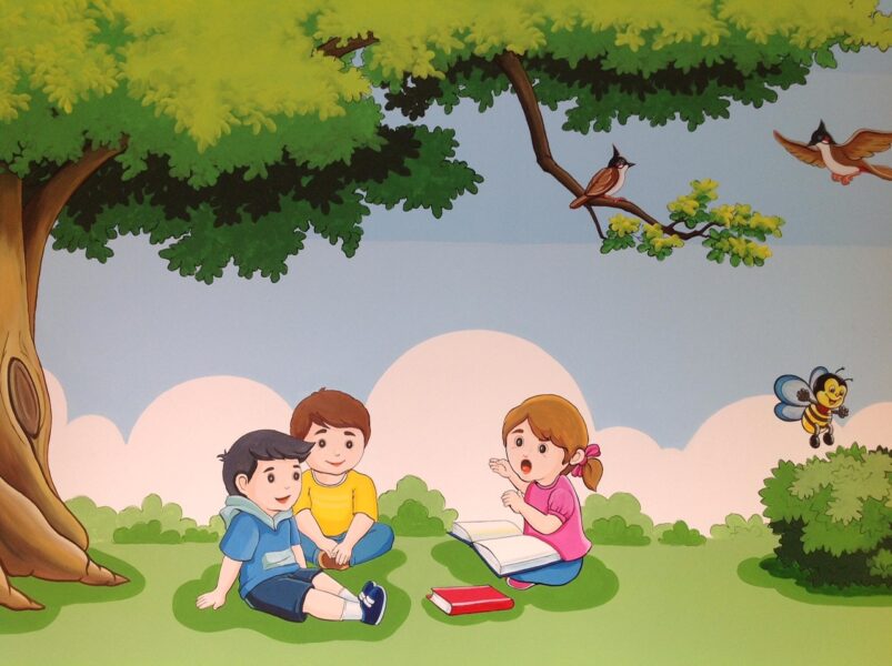 Tranh tường mầm non đẹp hình các bạn nhỏ ngồi đọc sách, chơi dưới gốc cây